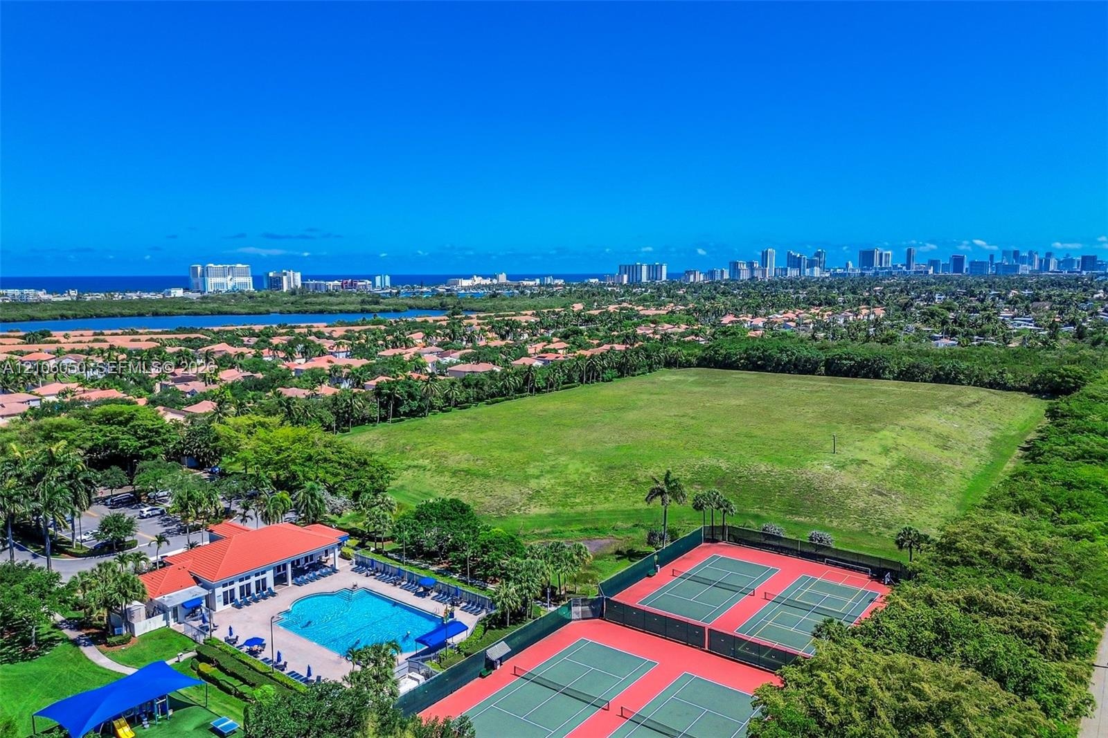 Aerial perspective featuring a clubhouse, an outdoor swimming pool with a surrounding paved deck, and two hard-surface tennis courts