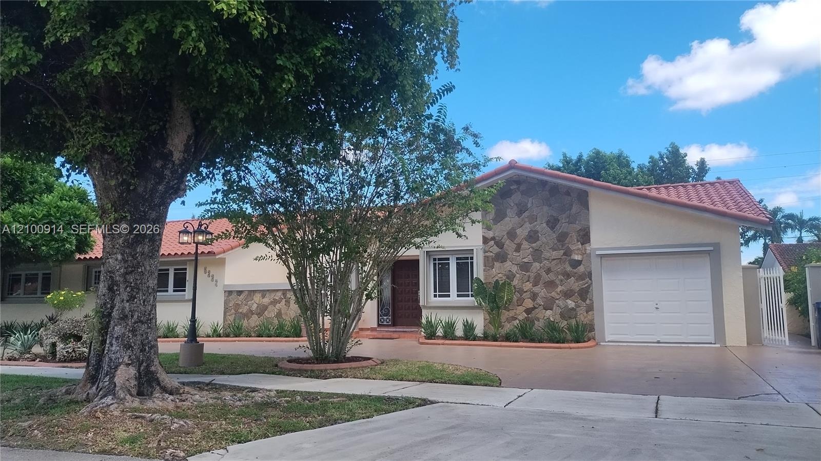 Single-story residence featuring a tile roof, light-colored exterior facade with stone accent detailing, attached garage, and mature landscape