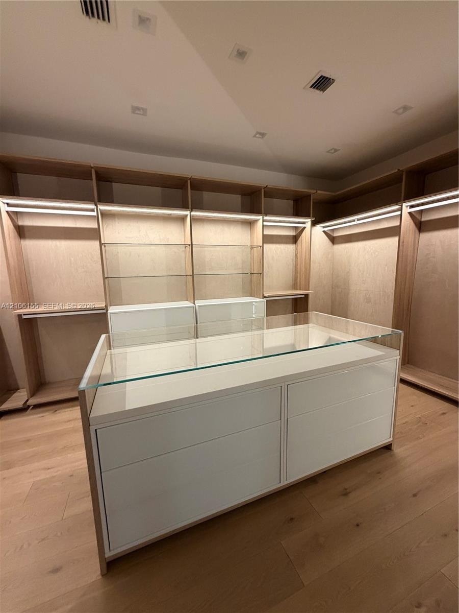 Custom Women’s primary closet , long mirror ,built-in wood cabinetry featuring integrated lighting and glass shelving, centered by a matching island w