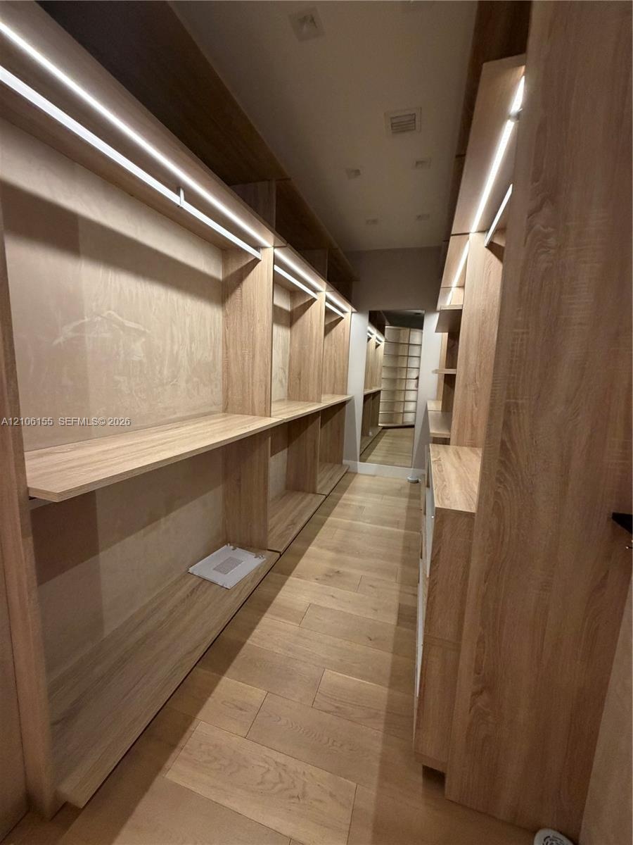 Custom Men’s Primary closet w storage system featuring light wood-finish cabinetry, integrated linear LED shelving illumination, open-concept shelving